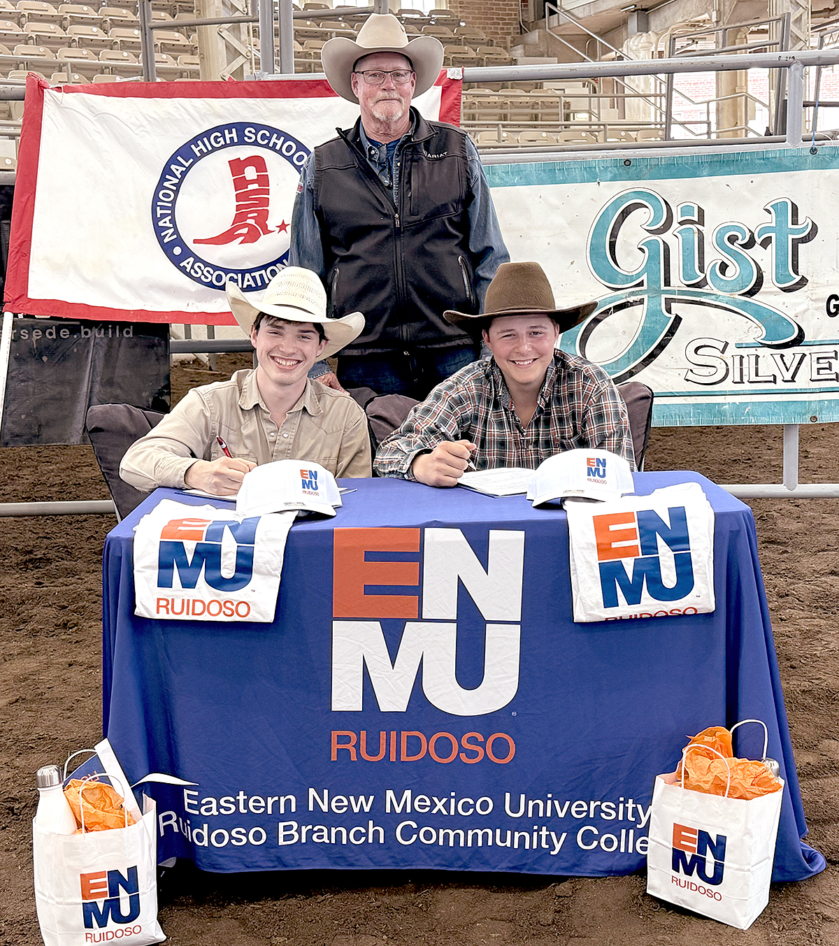 Photo of Walker Hanafin and Eli Foster sitting at table with ENMU-Ruidoso branded items, signing letters of intent to college rodeo at ENMU-Ruidoso with Illinois High School Rodeo National Director Bill Berger standing behind them.