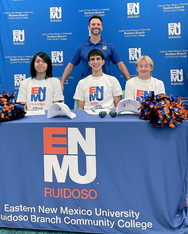 Photo of Gavin Castillo, Simon Vasquez and Jack Wisdom seated at table, all wearing ENMU-Ruidoso Cross Country t-shirts. Standing behind them is ENMU-Ruidoso Cross Country Head Coach Micah Bray.